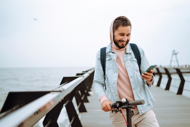 Junge Mann mit Telefon fährt mit einem elektrischen Roller auf einem Pier in der Nähe des Meeres Ökologisches Transportkonzept