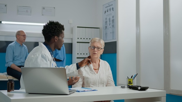 Junge Medizinerin, die kranke ältere Frau mit Krankheit in der Arztpraxis berät. Arzt, der einen alten Patienten am Schreibtisch untersucht, während eine Krankenschwester und ein älterer Mann auf dem Bett im Hintergrund sprechen