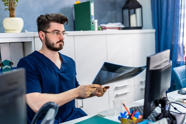 Junge medizinische Arbeitskraft, die am Schreibtisch sitzt. Bärtiger Arzt mit Brille, der Röntgen vor dem Monitor analysiert. Medizin-Konzept.