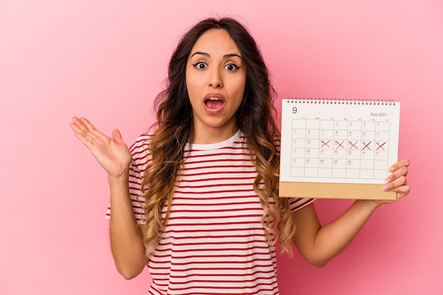 Junge mexikanische Frau, die einen Kalender einzeln auf rosafarbenem Hintergrund hält überrascht und schockiert.