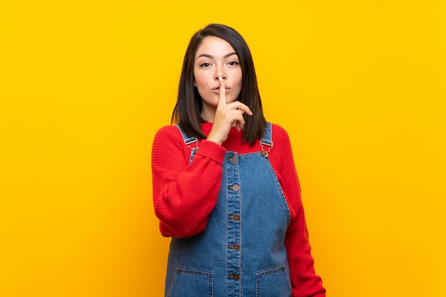 Junge mexikanische Frau mit Overall über der gelben Wand, die ein Zeichen der Ruhegeste einsetzt Finger in Mund zeigt