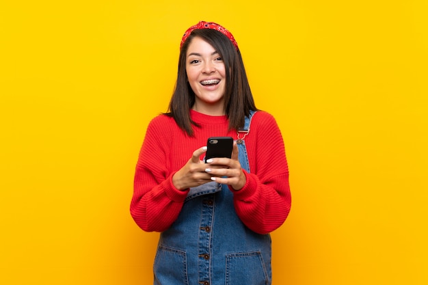 Junge mexikanische Frau mit Overall über der gelben Wand, die eine Mitteilung mit dem Mobile sendet