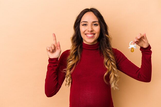 Junge mexikanische schwangere Frau, die Schnuller lokalisiert auf beige Wand zeigt, die Nummer eins mit Finger zeigt.