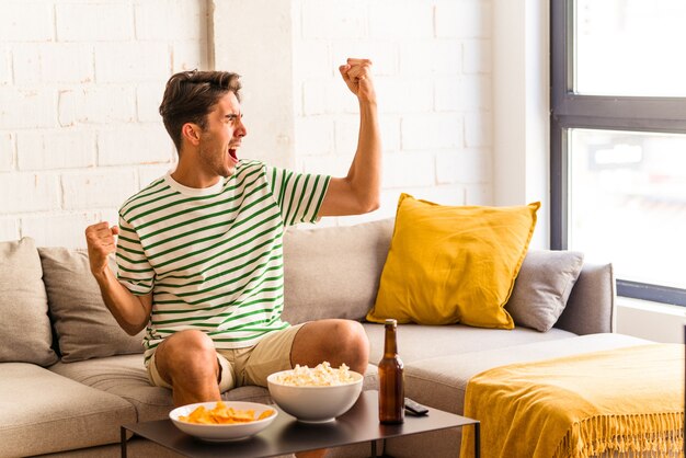 Junge Mischlinge, die Popcorn essen, sitzen auf dem Sofa und heben die Faust nach einem Sieg, Gewinnerkonzept.