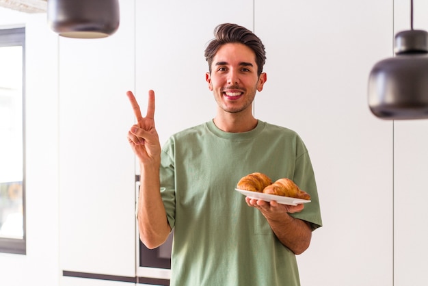 Junge Mischlinge essen Croissant in einer Küche am Morgen und zeigen Nummer zwei mit den Fingern.