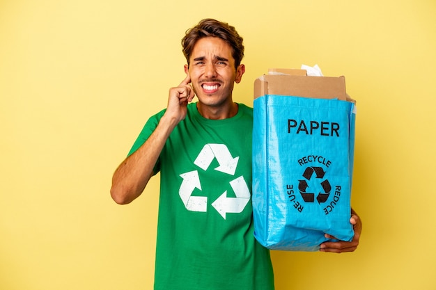 Junge Mischlinge Mann Recyclingpapier isoliert auf gelbem Hintergrund für Ohren mit Händen.