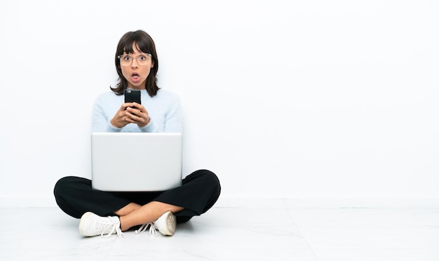 Junge Mischlingsfrau, die auf dem Boden sitzt, mit Laptop isoliert auf weißem Hintergrund, überrascht und Senden einer Nachricht