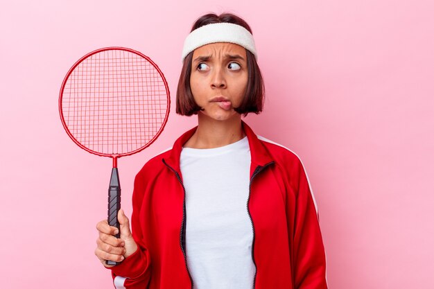 Junge Mischlingsfrau, die Badminton spielt, isoliert auf rosa Hintergrund verwirrt, fühlt sich zweifelhaft und unsicher.