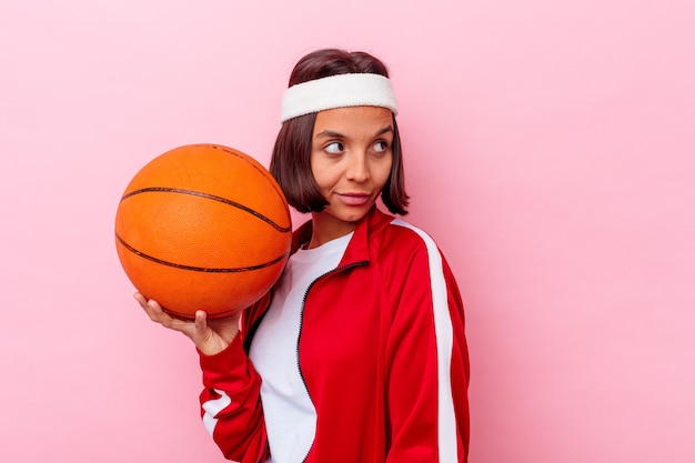 Junge Mischlingsfrau, die Basketball spielt, der auf rosa Wand lokalisiert aussieht, schaut beiseite lächelnd, fröhlich und angenehm.