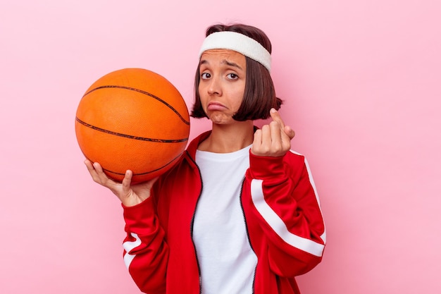 Junge Mischlingsfrau, die Basketball spielt, lokalisiert auf rosa Wand, die mit Finger auf Sie zeigt, als ob die Einladung näher kommt.