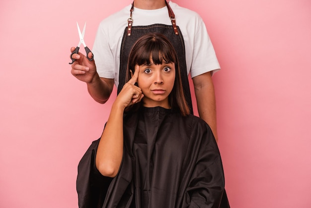 Foto junge mischlingsfrau, die beim friseur einen haarschnitt bekommt, isoliert auf rosafarbenem hintergrund, zeigt auf die schläfe mit fingerdenken, das sich auf eine aufgabe konzentriert