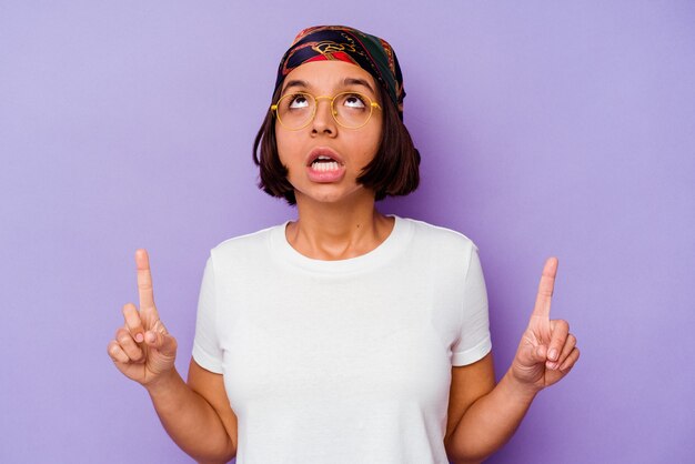 Junge Mischlingsfrau, die ein Bandana trägt, das auf violettem Hintergrund isoliert ist und mit geöffnetem Mund nach oben zeigt.