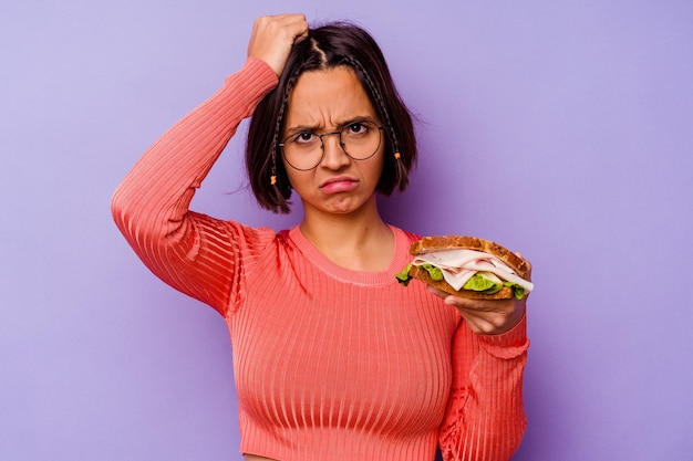 Junge Mischlingsfrau, die ein Sandwich einzeln auf violettem Hintergrund hält und schockiert ist, hat sich an ein wichtiges Treffen erinnert.