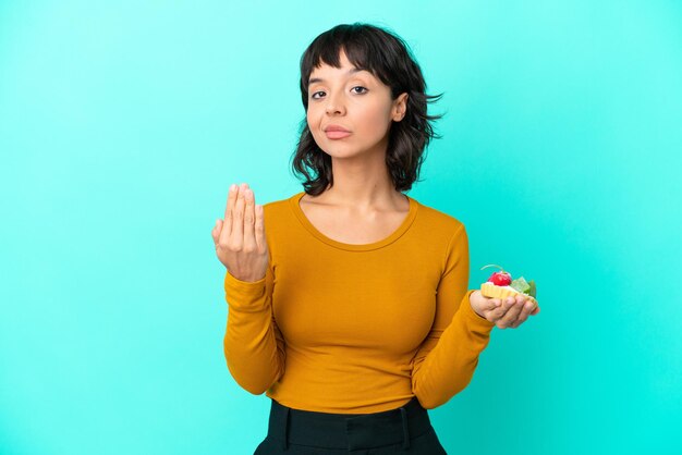 Junge Mischlingsfrau, die ein Törtchen hält, das auf blauem Hintergrund isoliert ist und einlädt, mit der Hand zu kommen. Glücklich, dass Sie gekommen sind