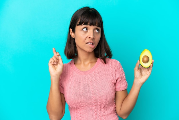Junge Mischlingsfrau, die eine Avocado auf blauem Hintergrund mit gekreuzten Fingern hält und das Beste wünscht