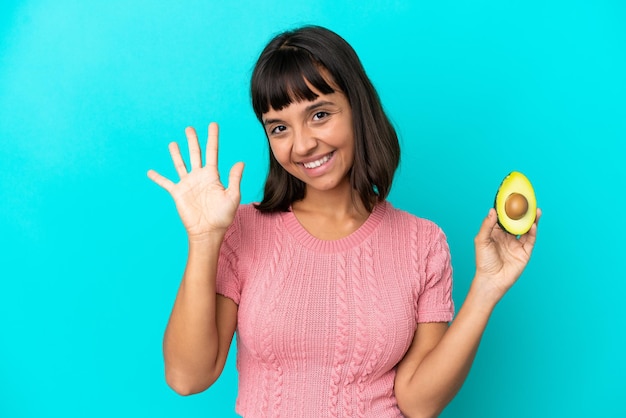 Junge Mischlingsfrau, die eine Avocado hält, die auf blauem Hintergrund isoliert ist und fünf mit den Fingern zählt