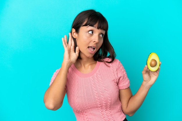 Junge Mischlingsfrau, die eine Avocado isoliert auf blauem Hintergrund hält und etwas hört, indem sie die Hand auf das Ohr legt