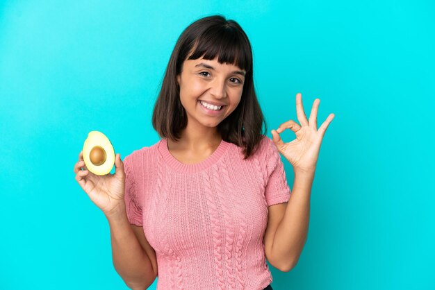 Junge Mischlingsfrau, die eine Avocado isoliert auf blauem Hintergrund hält und mit den Fingern ein Ok-Zeichen zeigt