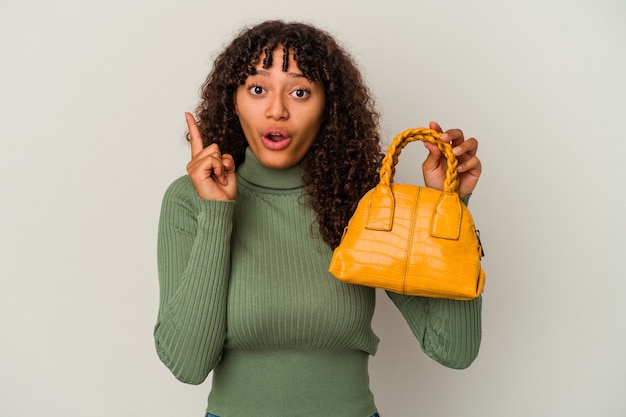 Junge Mischlingsfrau, die eine Handtasche lokalisiert auf weißem Hintergrund mit einer Idee, Inspirationskonzept hält.