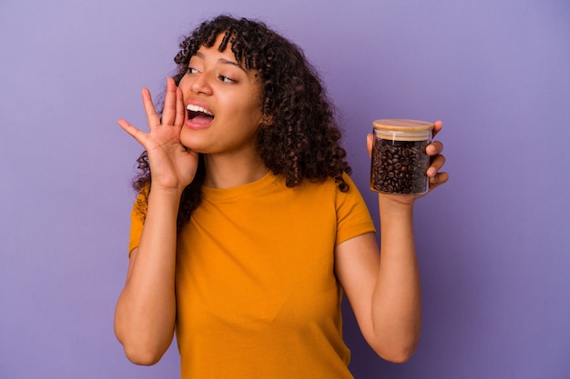 Junge mischlingsfrau, die eine kaffeebohnenflasche lokalisiert auf violettem hintergrund hält, die palme nahe geöffnetem mund schreit und hält.