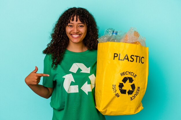 Junge Mischlingsfrau, die eine recycelte Plastiktüte einzeln auf blauem Hintergrund hält, die mit der Hand auf einen Hemdkopierraum zeigt, stolz und selbstbewusst