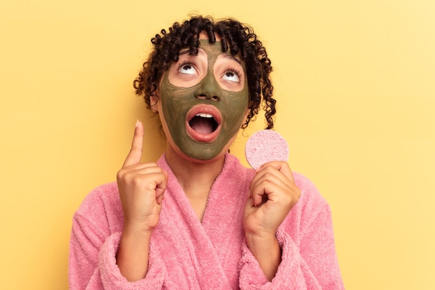 Junge Mischlingsfrau, die einen Bademantel trägt, der einen Make-up-Entfernerschwamm hält, der auf gelbem Hintergrund isoliert ist und mit geöffnetem Mund nach oben zeigt.