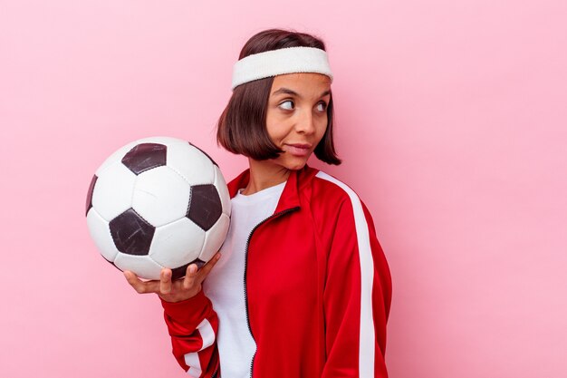 Junge Mischlingsfrau, die Fußball spielt, der auf rosa Wand lokalisiert aussieht, schaut beiseite lächelnd, fröhlich und angenehm.