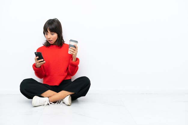 Junge Mischlingsfrau, die isoliert auf weißem Hintergrund auf dem Boden sitzt und Kaffee zum Mitnehmen und ein Handy hält