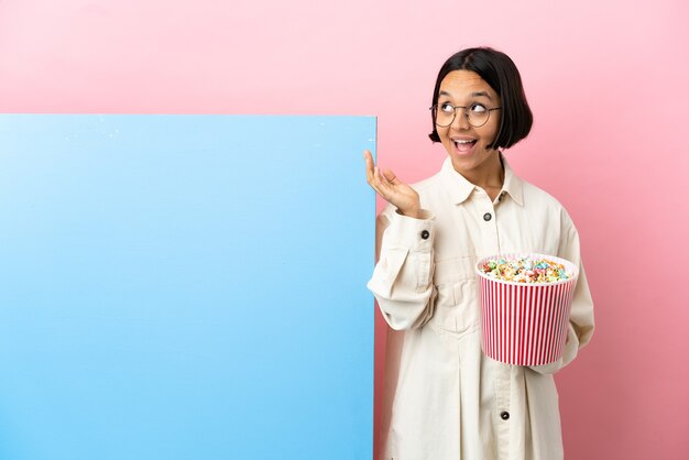 Junge Mischlingsfrau, die Popcorn mit einem großen Banner über isoliertem Hintergrund mit überraschendem Gesichtsausdruck hält