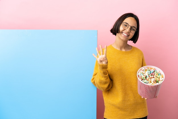 Junge Mischlingsfrau, die Popcorns mit einem großen Banner über lokalisiertem Hintergrund glücklich hält und vier mit den Fingern zählt