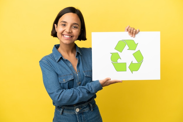 Junge Mischlingsfrau isoliert auf gelbem Hintergrund, die ein Plakat mit Recycling-Symbol mit glücklichem Ausdruck hält