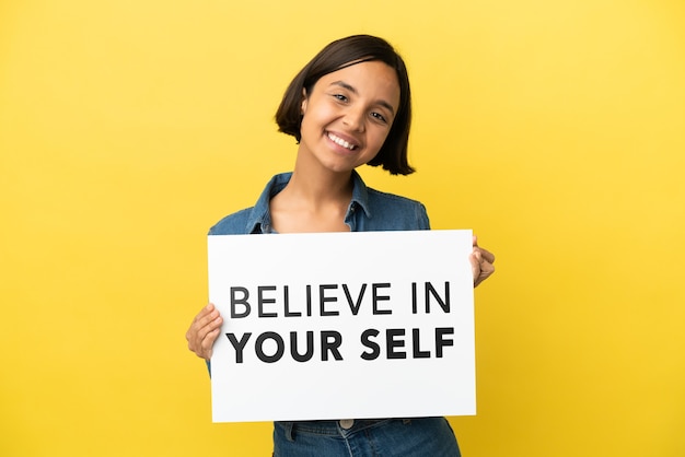 Junge Mischlingsfrau lokalisiert auf gelbem Hintergrund, der ein Plakat mit Text glaubt an Ihr Selbst mit glücklichem Ausdruck hält
