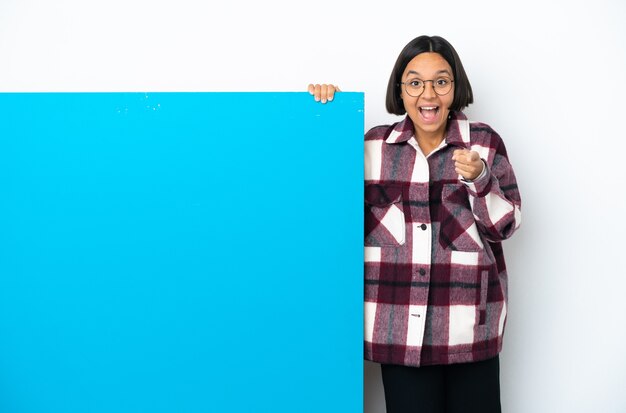 Junge Mischlingsfrau mit einem großen blauen Plakat lokalisiert auf weißem Hintergrund überrascht und nach vorne zeigend