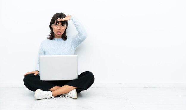 Junge Mischlingsfrau sitzt auf dem Boden mit Laptop isoliert auf weißem Hintergrund und schaut weit weg mit der Hand, um etwas zu sehen