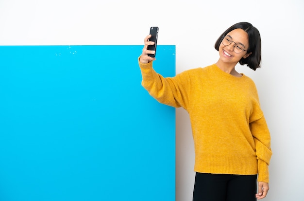 Junge Mischrassenfrau mit einem großen blauen Plakat lokalisiert auf weißem Hintergrund, der ein Selfie macht