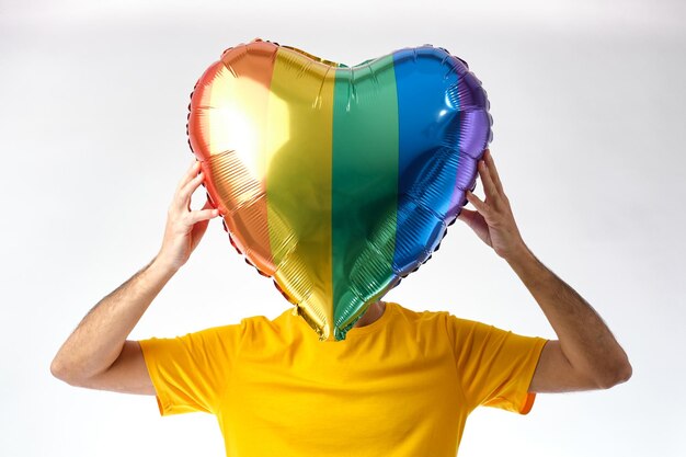 Junge mit brauner Haut in einem gelben T-Shirt, die einen herzförmigen Ballon mit den Farben der Gay-Pride-Flagge vor ihrem Gesicht auf weißem Hintergrund hält