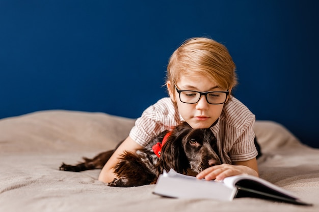 Junge mit Brille und mit seinem Hund liegt auf dem Bett und liest ein großes Buch.