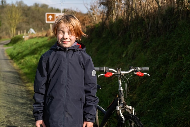 Junge mit Fahrrad auf dem Land Junge im Alter von neun oder zehn Jahren steht auf grünem Gras in der Nähe der Straße Es gibt ein Fahrrad in der Nähe Aktives Freizeitradfahren