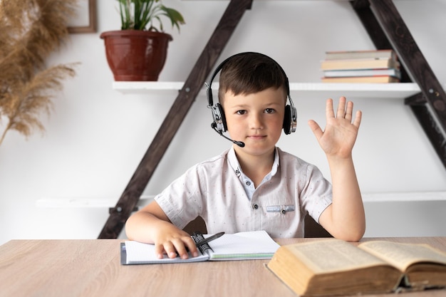 Junge mit Kopfhörern sitzt am Tisch und winkt mit der Hand in die Kamera Online-Unterricht oder Nachhilfelehrer für zu Hause unterrichtetes Kind Grundschüler mit drahtlosem Headset