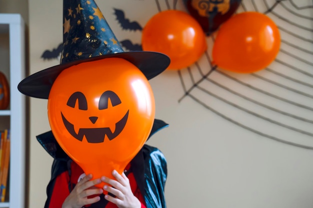Junge mit schwarzem Umhang und Hexenhut hält Luftballon mit Kürbis-Jack im Gesicht. Halloween-Feier