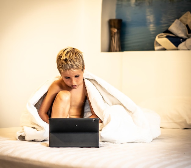 Junge mit Tablet-Uhr-Cartoon unter Decke morgens oder abends im Bett, wenn die Eltern beschäftigt sind oder schlafen Abhängigkeit vom Fernsehen Schwierigkeiten bei der Kontrolle der Inhalte von Kindern im Internet-Konzept