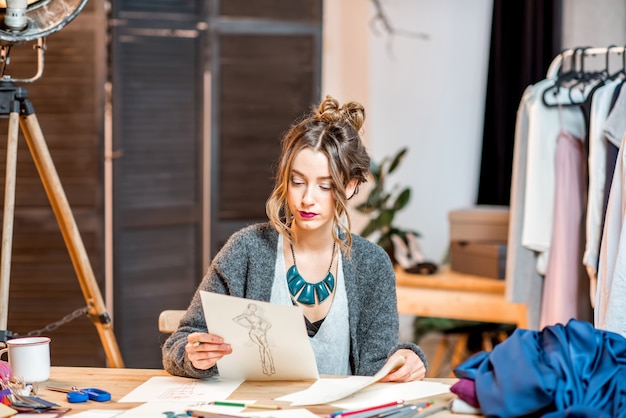 Foto junge modedesignerin, die zeichnungen der kleidung skizziert, die im schönen büro mit verschiedenen schneiderwerkzeugen sitzt