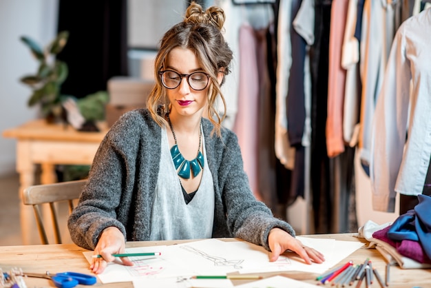 Junge Modedesignerin, die Zeichnungen der Kleidung skizziert, die im schönen Büro mit verschiedenen Schneiderwerkzeugen sitzt