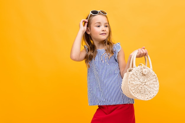 Junge Modefan mit einer runden Nachtigallhandtasche und einer Brille auf ihrem Kopf, die auf einer leuchtend gelben Wand aufwirft