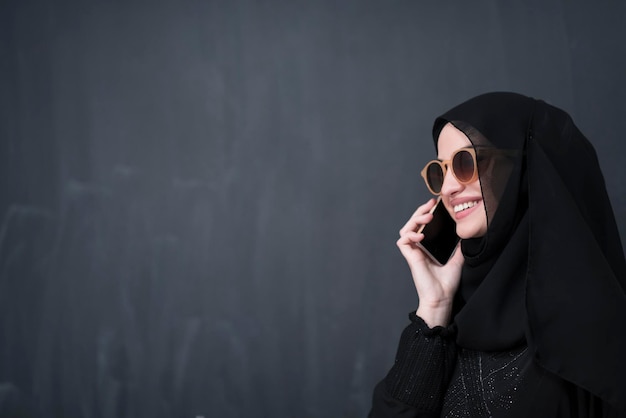 Junge moderne muslimische Geschäftsfrau mit Smartphones mit Sonnenbrille und Hijab-Kleidung vor einer schwarzen Tafel.