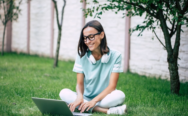 Junge moderne schöne glückliche selbstbewusste Frau in Kopfhörern und Brille arbeitet an einem Laptop, während auf dem Rasen sitzend.