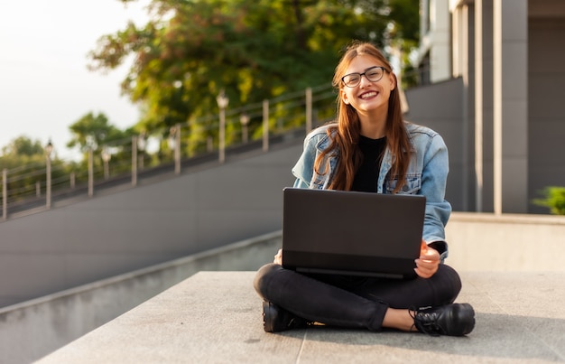 Junge moderne Studentin in einer Jeansjacke, die auf der Treppe mit Laptop sitzt. Fernunterricht. Modernes Jugendkonzept.