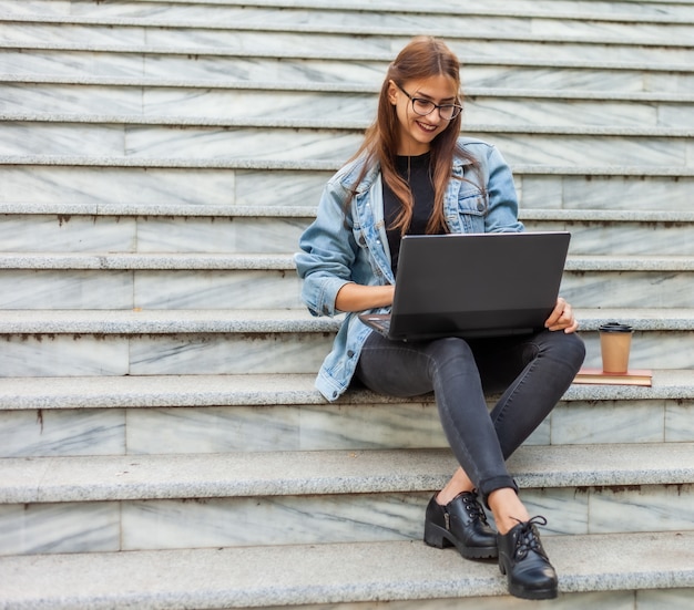 Junge moderne Studentin in einer Jeansjacke, die auf der Treppe sitzt und Video auf Laptop betrachtet. Fernunterricht. Modernes Jugendkonzept.