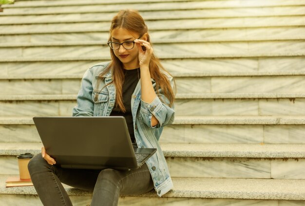 Junge moderne Studentin in einer Jeansjacke, die mit Laptop auf der Treppe sitzt. Video schauen. Fernunterricht. Modernes Jugendkonzept.