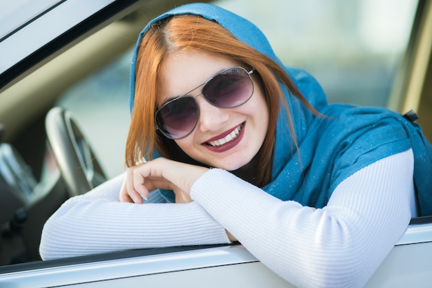 Junge modische Fahrerin im Schal und in der Sonnenbrille, die aus dem vorderen Fenster des Autos schauen.